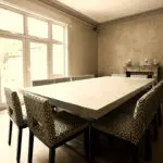 A spacious dining room with a large white table and patterned chairs, illuminated by natural light from a window.