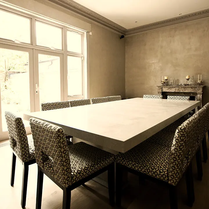 A spacious dining room with a large white table and patterned chairs, illuminated by natural light from a window.