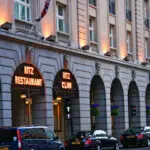 The image shows the exterior of a building at dusk with illuminated signs that read "ritz restaurant" and "ritz club." there are arched entryways, several lit windows, and flower boxes with purple flowers. cars are parked out front, suggesting this is a street view in a city.