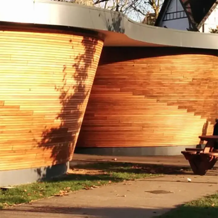 Curved wooden wall with shadows cast by the evening sun, adjacent to a paved walkway with a park bench in the background.