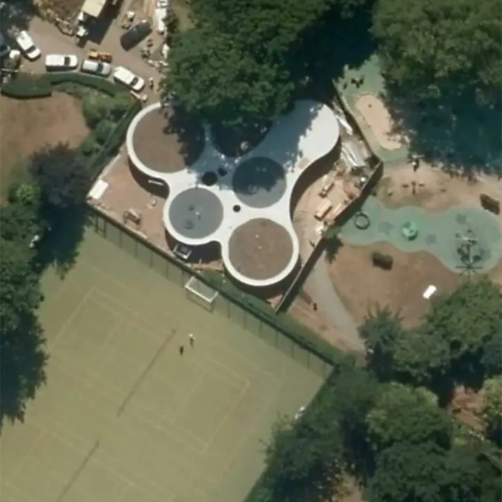 Aerial view of a unique building resembling a cloverleaf pattern next to a tennis court and outdoor pool surrounded by trees.