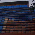 A low-angle view of a modern building with reflective glass windows and metal facade elements at dusk, framed by foliage in the foreground.