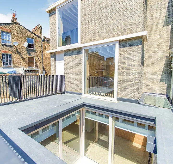 A modern rooftop terrace with glass balustrades and a walkable glass skylight providing natural light to the space below, surrounded by traditional brick buildings.