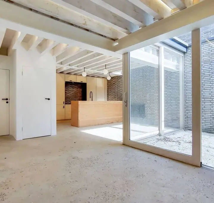 An unfinished room with exposed ceiling beams, large glass doors leading to a courtyard, and construction materials on the floor.