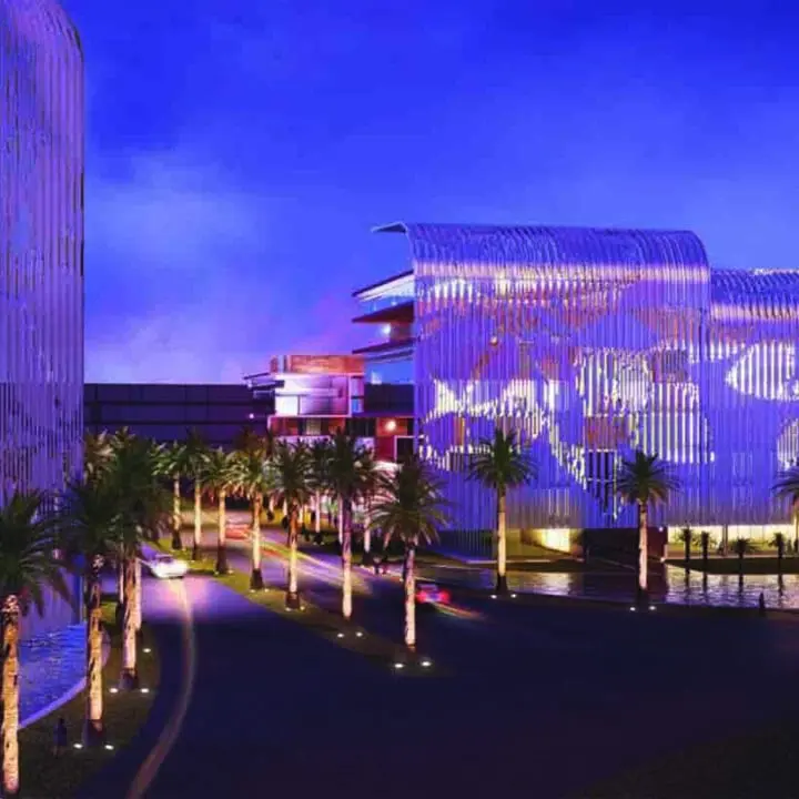 Modern buildings illuminated with blue lights at night, featuring a palm-tree-lined street and reflections on the glass facades.