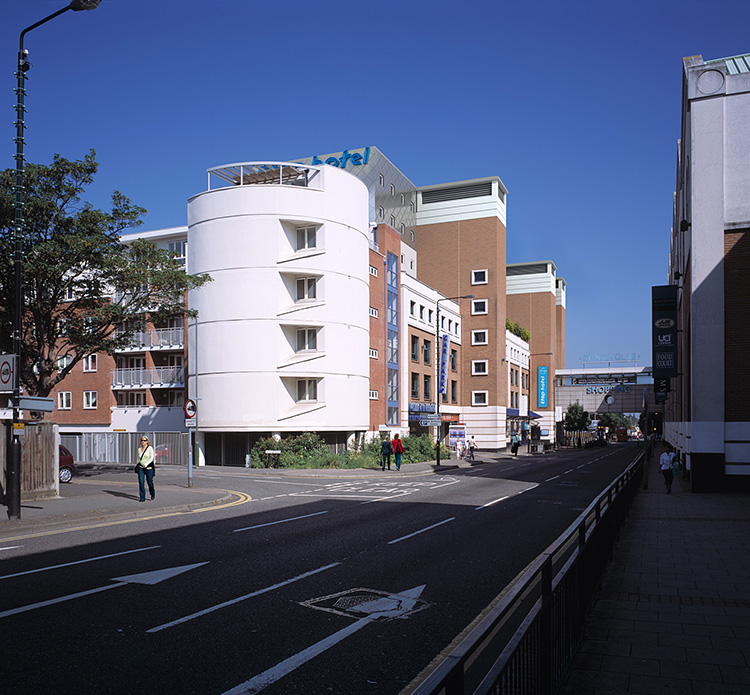 A curved white building stands at a street corner on a sunny day, with pedestrians nearby and various other buildings in the background.